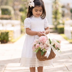 A young girl in a white dress stands in a sunlit outdoor walkway between columns, holding a wicker basket filled with pink flowers and tied with a white ribbon.