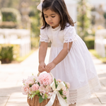 A young girl in a white dress stands in a garden walkway holding a wicker basket filled with pink flowers. The background features blurred stone columns and greenery.