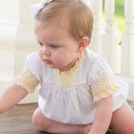 A baby girl wearing a white and yellow dress crawls on a wooden porch floor, reaching toward a small green leaf. She has a white bow in her hair, with a white railing and green garden background.