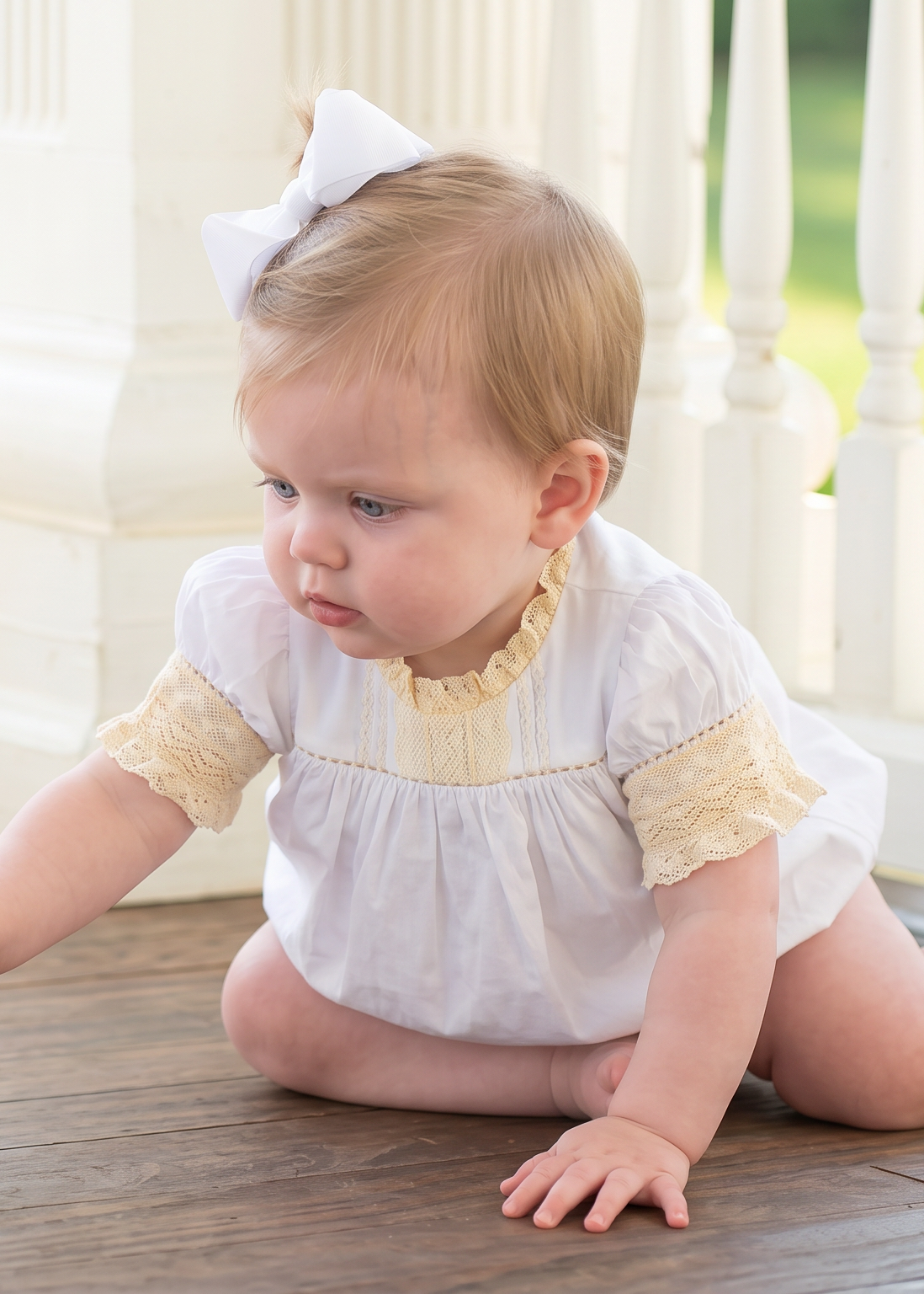 A baby girl wearing a white and yellow dress crawls on a wooden porch floor, reaching toward a small green leaf. She has a white bow in her hair, with a white railing and green garden background.