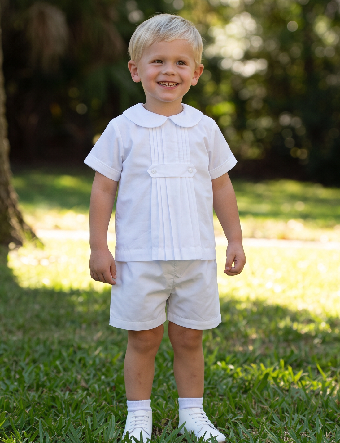 A young toddler boy standing on a grassy lawn in front of a large tree, wearing a white short-sleeve collared shirt, white shorts, and white socks.