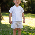 A young toddler boy standing on a grassy lawn in front of a large tree, wearing a white short-sleeve collared shirt, white shorts, and white socks.