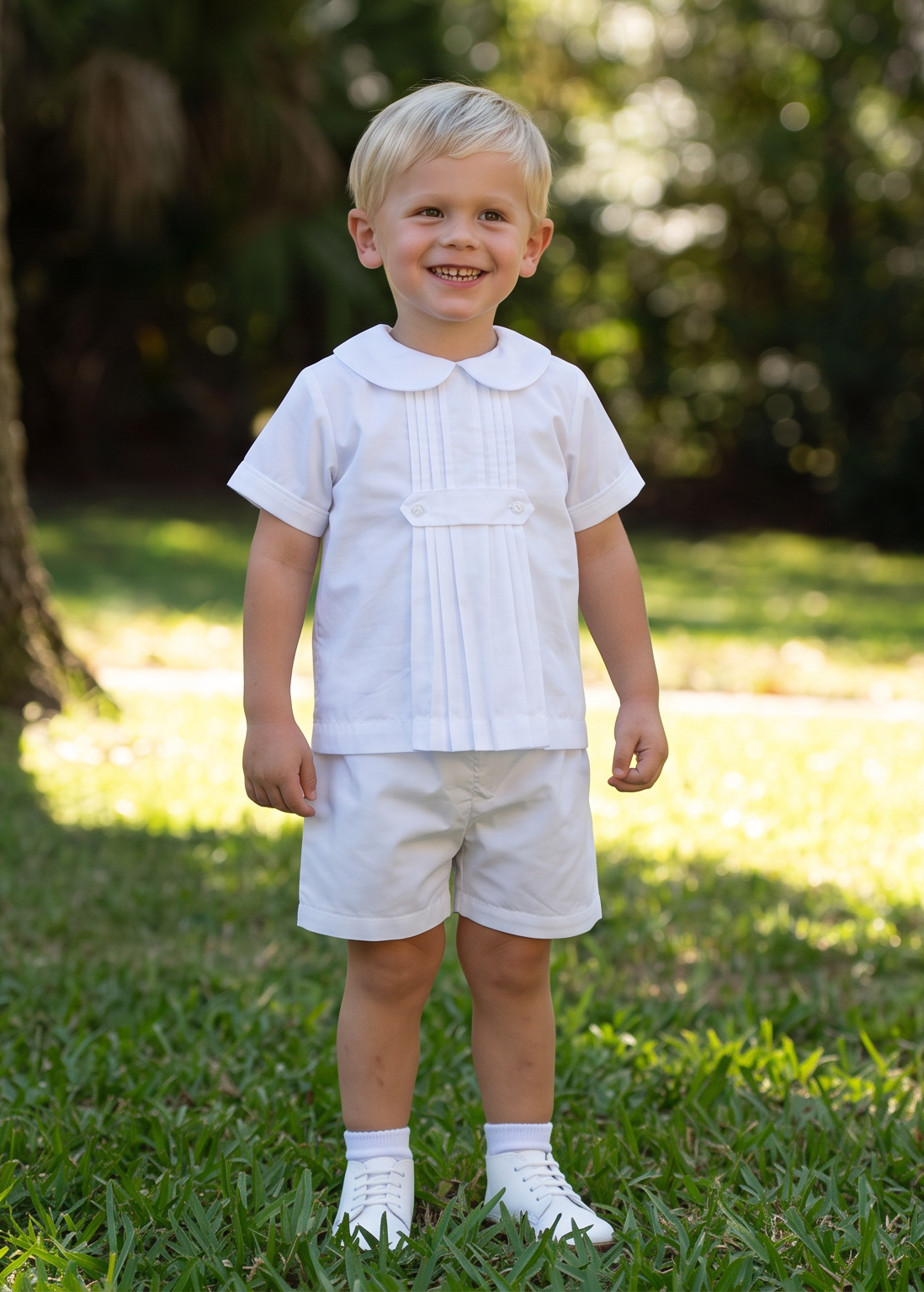 A young toddler boy standing on a grassy lawn in front of a large tree, wearing a white short-sleeve collared shirt, white shorts, and white socks.
