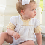 A baby girl sitting on a wooden porch wearing a white smocked bodysuit with yellow lace sleeves and a white bow in her hair, looking to the side.