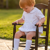 A toddler with strawberry-blonde hair sits in a miniature dark wooden rocking chair on a stone patio in a sunny backyard. The child is wearing a white short-sleeve dress, white socks, and white shoes.