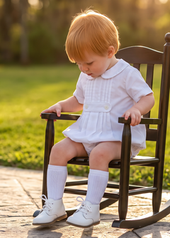 A toddler with strawberry-blonde hair sits in a miniature dark wooden rocking chair on a stone patio in a sunny backyard. The child is wearing a white short-sleeve dress, white socks, and white shoes.