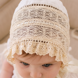 A baby wearing a cream-colored lace bonnet with ribbon ties, sitting on a beige upholstered chair.