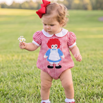 A toddler girl wearing a red and white gingham raggedy doll romper stands in a grassy park. The romper features a white Peter Pan collar and an embroidered character of a girl with red yarn hair, a red dress, and a blue apron.