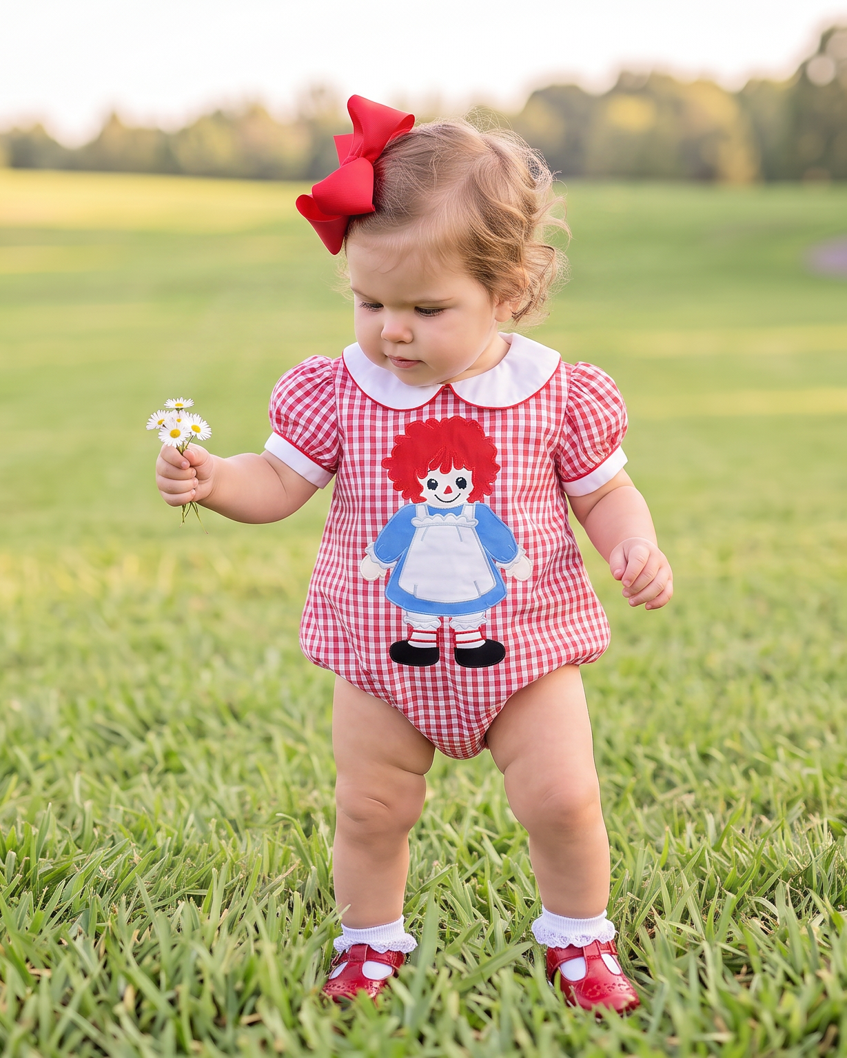 A toddler girl wearing a red and white gingham raggedy doll romper stands in a grassy park. The romper features a white Peter Pan collar and an embroidered character of a girl with red yarn hair, a red dress, and a blue apron.