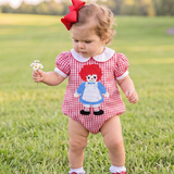 A toddler girl wearing a red and white gingham raggedy doll romper stands in a grassy park. The romper features a white Peter Pan collar and an embroidered character of a girl with red yarn hair, a red dress, and a blue apron.