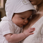 A baby in a white baptism gown and matching bonnet held by a woman in a white lace dress inside a church, with a stone baptismal font in the background.