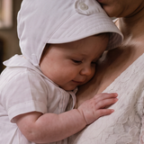 A baby in a white baptism gown and matching bonnet held by a woman in a white lace dress inside a church, with a stone baptismal font in the background.