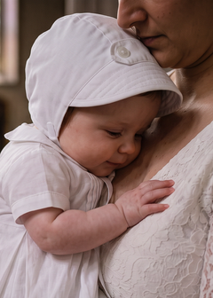 A baby in a white baptism gown and matching bonnet held by a woman in a white lace dress inside a church, with a stone baptismal font in the background.