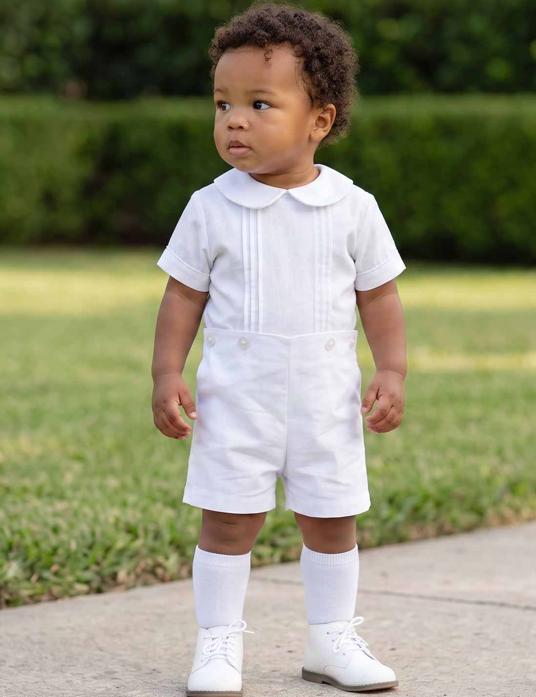 A toddler standing on a concrete path in a grassy yard, wearing a white short-sleeve collared romper with white knee-length socks and white shoes.