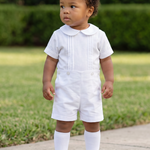 A toddler standing on a concrete path in a grassy yard, wearing a white short-sleeve collared romper with white knee-length socks and white shoes.