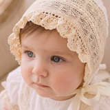 A baby wearing a cream lace bonnet and matching cream dress, sitting on a beige upholstered armchair in a softly lit indoor setting.