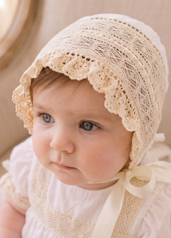 A baby wearing a cream lace bonnet and matching cream dress, sitting on a beige upholstered armchair in a softly lit indoor setting.