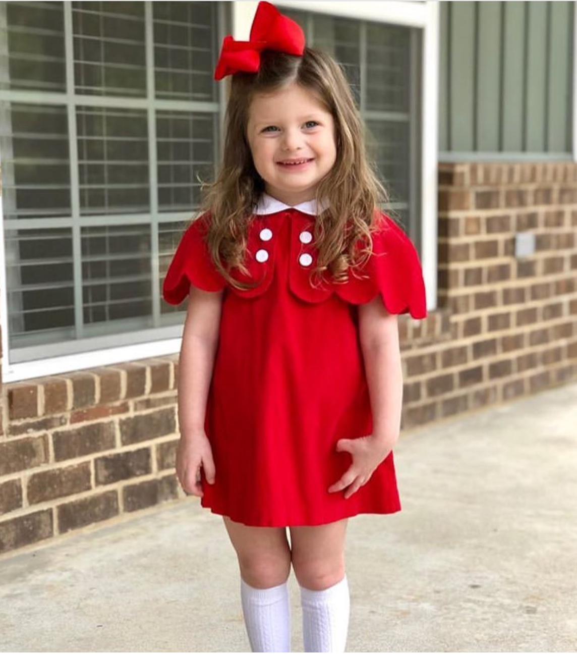 Young girl in a red dress with white buttons standing in front of a brick building.
