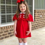 Young girl in a red dress with white buttons standing in front of a brick building.