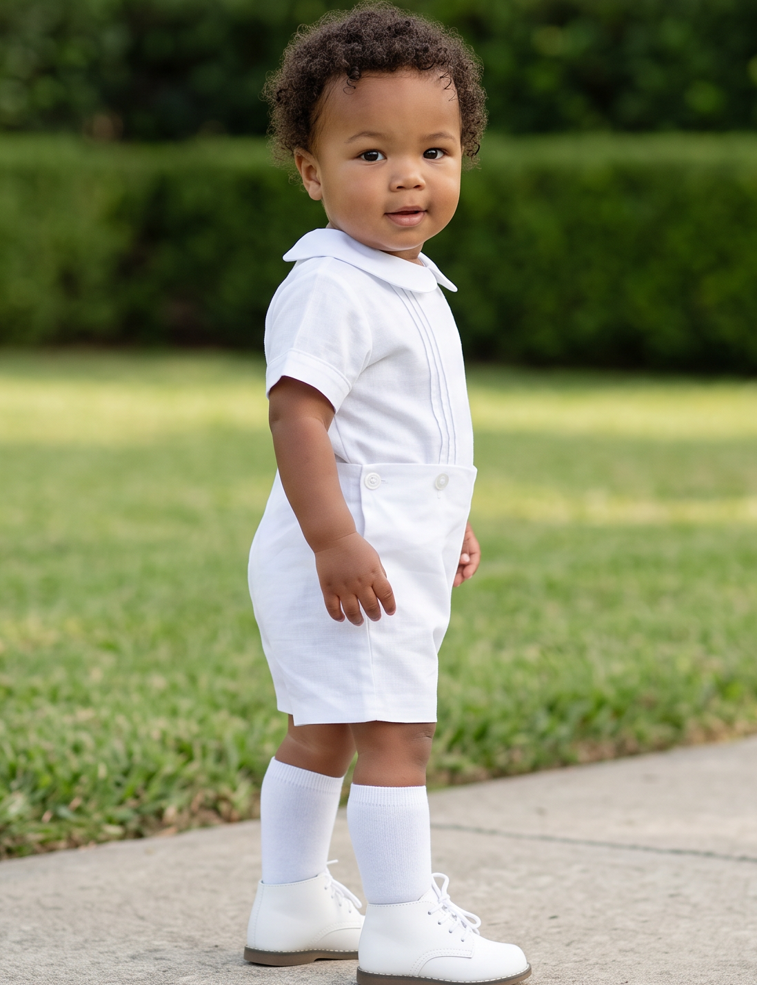 A toddler stands on a concrete path in a grassy yard wearing a white short-sleeve polo shirt, white shorts, white socks, and white shoes.