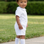 A toddler stands on a concrete path in a grassy yard wearing a white short-sleeve polo shirt, white shorts, white socks, and white shoes.