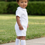 A toddler stands on a concrete path in a grassy yard wearing a white short-sleeve polo shirt, white shorts, white socks, and white shoes.