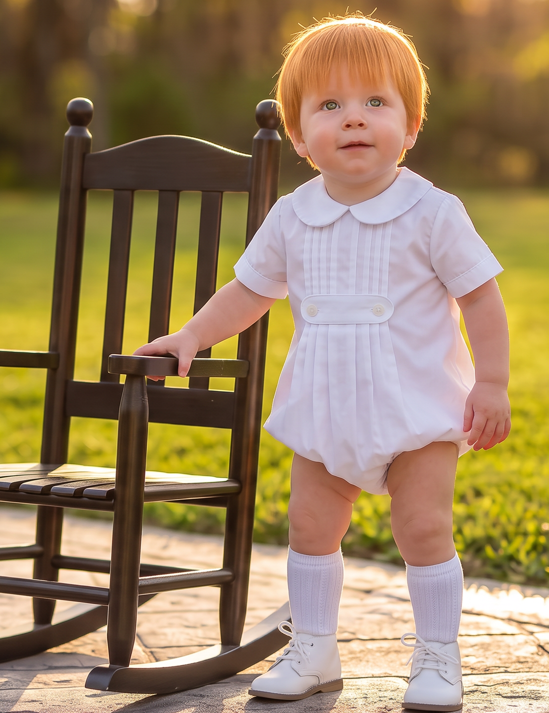A toddler with red hair stands on a stone path in a park next to a dark wooden rocking chair, wearing a white short-sleeve romper, white socks, and white shoes.