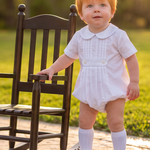 A toddler with red hair stands on a stone path in a park next to a dark wooden rocking chair, wearing a white short-sleeve romper, white socks, and white shoes.