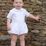 A toddler standing barefoot on stone steps outdoors, wearing a white short-sleeve romper dress with a Peter Pan collar.
