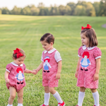 Three young children wearing matching red-and-white gingham dresses with a rag doll graphic stand on a grassy park path. The dresses have white collars and cuffs, white socks, and red shoes, and each child wears a red bow headband.