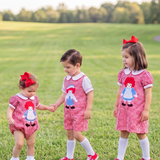 Three young children wearing matching red-and-white gingham dresses with a rag doll graphic stand on a grassy park path. The dresses have white collars and cuffs, white socks, and red shoes, and each child wears a red bow headband.