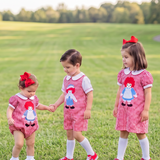 Three young children wearing matching red-and-white gingham dresses with a rag doll graphic stand on a grassy park path. The dresses have white collars and cuffs, white socks, and red shoes, and each child wears a red bow headband.