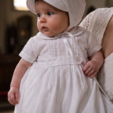 A baby boy in a white first communion dress and bonnet stands on a church porch being held by a woman in a white lace dress. A stone baptismal font and stained glass windows are visible in the softly lit background.
