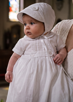 A baby boy in a white first communion dress and bonnet stands on a church porch being held by a woman in a white lace dress. A stone baptismal font and stained glass windows are visible in the softly lit background.