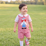 A toddler walking in a grassy park wearing a red and white gingham overall romper with a Raggedy Ann graphic on the chest, a white t-shirt, white socks, and red sneakers.
