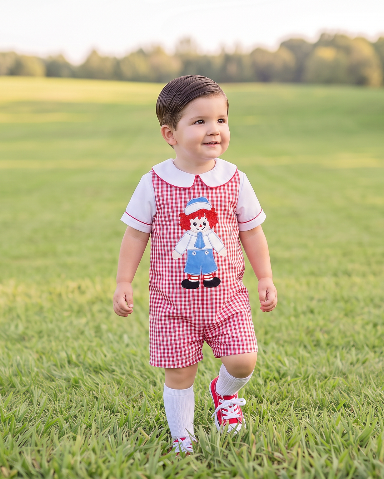 A toddler walking in a grassy park wearing a red and white gingham overall romper with a Raggedy Ann graphic on the chest, a white t-shirt, white socks, and red sneakers.