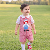 A toddler walking in a grassy park wearing a red and white gingham overall romper with a Raggedy Ann graphic on the chest, a white t-shirt, white socks, and red sneakers.