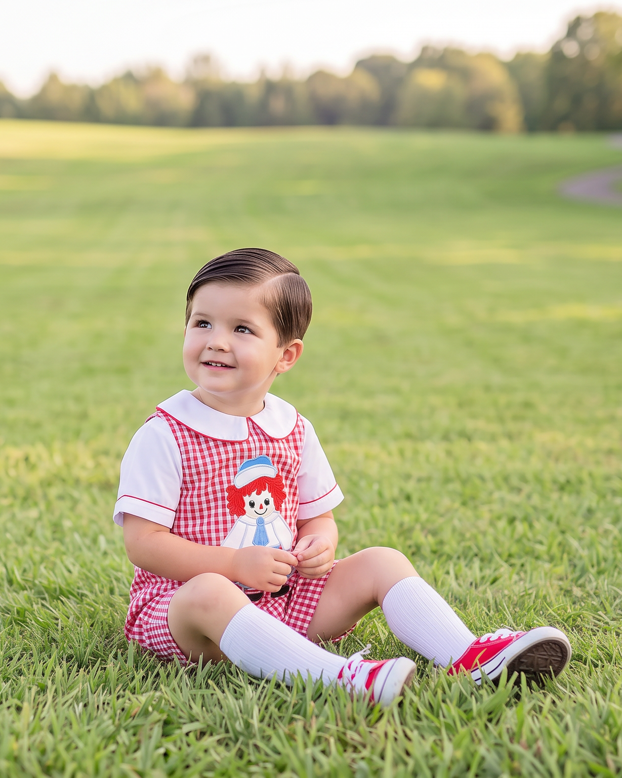 A toddler sitting on a green grassy field, wearing a red and white gingham dress with a red hat patch, white socks, and red-and-white sneakers.