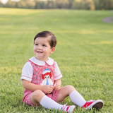 A toddler sitting on a green grassy field, wearing a red and white gingham dress with a red hat patch, white socks, and red-and-white sneakers.