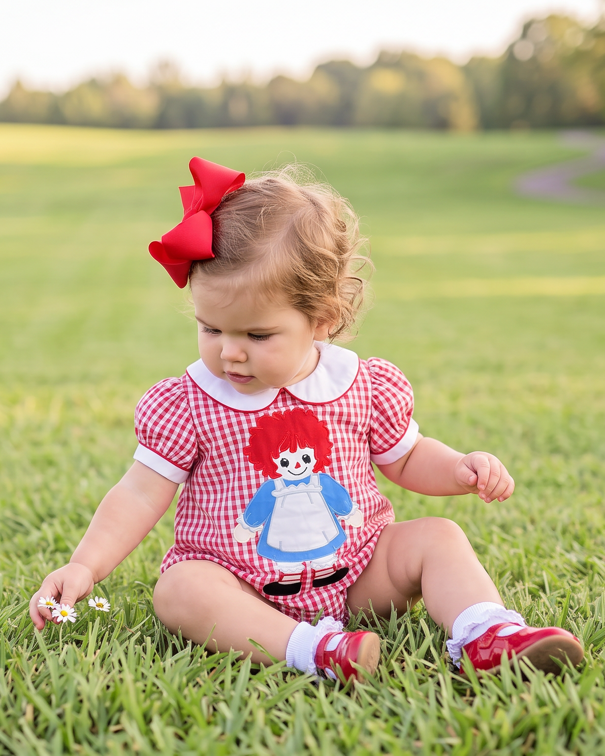 A toddler sitting on a green grassy field wearing a red and white gingham romper with a Raggedy Ann doll graphic on the front, a red bow in her hair, and red shoes.