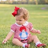 A toddler sitting on a green grassy field wearing a red and white gingham romper with a Raggedy Ann doll graphic on the front, a red bow in her hair, and red shoes.