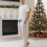 Man wearing Christmas-themed pajama pants standing in a living room with a decorated tree and fireplace.