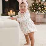 Baby in a floral dress standing in a festive living room with Christmas decorations.