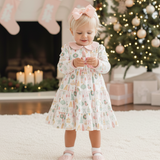 Young girl in a floral dress standing in front of a Christmas tree and fireplace.