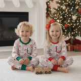 Two children in matching pajamas sitting on a floor decorated for Christmas with a tree and stockings.