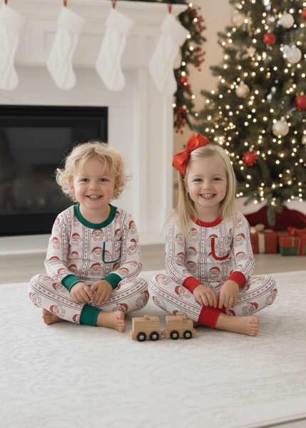 Two children in matching pajamas sitting on a floor decorated for Christmas with a tree and stockings.