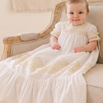A baby wearing a white first communion dress sits on a beige upholstered wooden armchair in a bright, neutral-toned room.