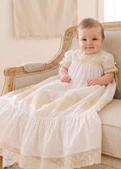 A baby wearing a white first communion dress sits on a beige upholstered wooden armchair in a bright, neutral-toned room.