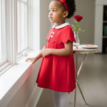 Young girl in a red dress standing by a window with a rose on a small table.