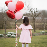 Child in a pink dress holding red and white heart balloons in a park.